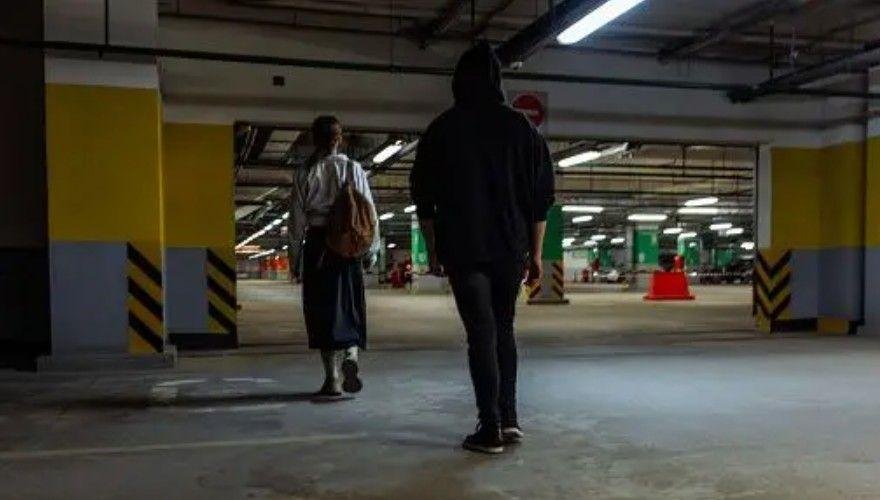 رجل يضع غطاء الرأس على رأسه يتعقب امرأة في موقف سيارات تحت الأرض خافت الإضاءة. 
